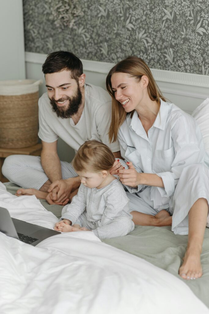 A young family of three enjoys watching content together on a laptop in a cozy bedroom setting.